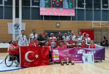 Türkiye Tekerlekli Sandalye Kadın Basketbol Takımı, spor salonunda Türk bayrakları ve Avrupa Şampiyonası pankartları eşliğinde önlerinde kupa ve madalyalarıyla birlikte gururla poz veriyor.