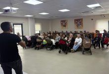 Görselde, bir salonda bir konuşmacının karşısında oturan kalabalık bir grup insan, çocuk arabaları ve çocuklarla birlikte dikkatle sunumu dinliyor; sağ tarafta ise bir kişi ayakta duruyor..