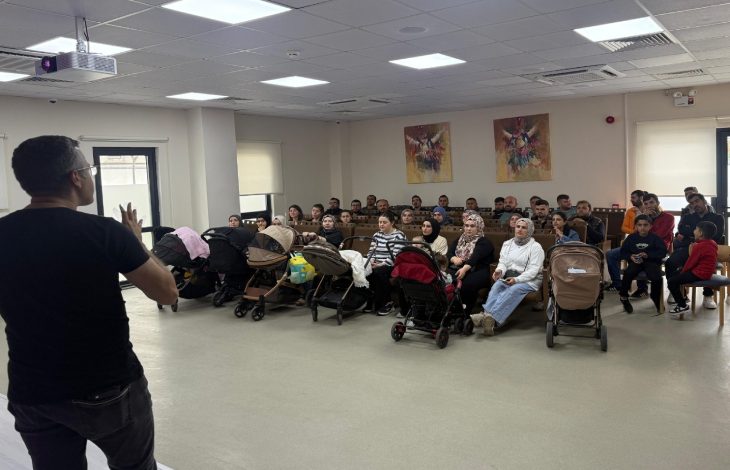 Görselde, bir salonda bir konuşmacının karşısında oturan kalabalık bir grup insan, çocuk arabaları ve çocuklarla birlikte dikkatle sunumu dinliyor; sağ tarafta ise bir kişi ayakta duruyor..
