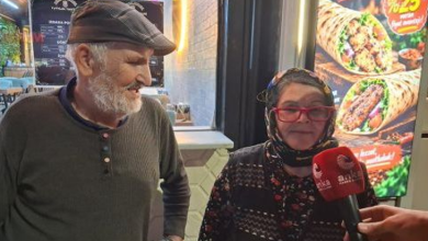 Görselde, bir sokak veya işletme önünde röportaj veren ileri yaşlarda iki kişi yer almaktadır. Sol tarafta kasketli ve sakallı bir erkek ayakta dururken, sağ tarafta ise başörtülü, kırmızı gözlüklü bir kadın ellerinde mikrofonla konuşmaktadır. Arka planda ise bir restorana ait yemek görselleri bulunan ışıklı bir pano görülmektedir.