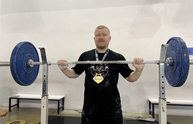 Görselde, spor salonunda halter barını kaldıran veya tutan, boynunda madalya takılı olan bir sporcu yer almaktadır.