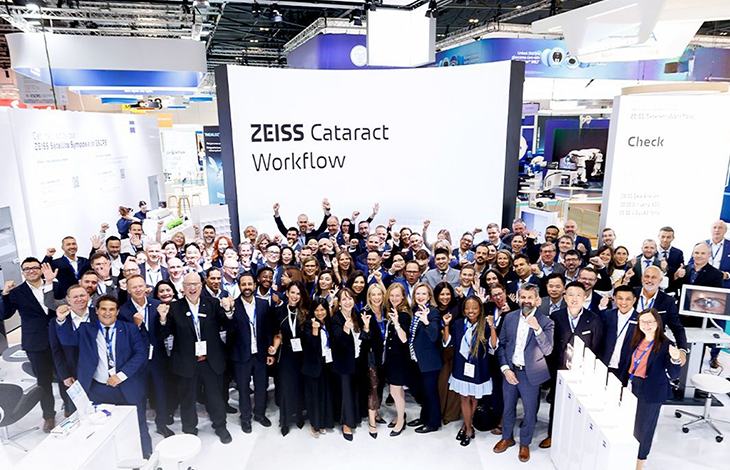 Kalabalık bir grup, aydınlık bir fuar alanında kurulan ZEISS Cataract Workflow standının önünde toplu halde poz veriyor. Katılımcıların çoğu iş kıyafeti ve yaka kartı taşıyor; ortam profesyonel bir kongre veya teknoloji etkinliği havasında. Arka planda modern tıbbi cihazlar ve parlak ışıklarla hazırlanmış sergi alanı görülüyor. Genel atmosfer, göz sağlığı ve katarakt teknolojilerine odaklanan kurumsal bir buluşmayı yansıtıyor.