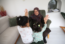 Görselin üst bölümünde, ev ortamında zeytin yeşili bir koltukta oturan başörtülü koruyucu anne ile önünde yer alan iki kız çocuğunun neşeyle el çırpma oyunu oynadığı anlar yer almaktadır. Kadın gülümseyerek çocuklara ellerini uzatırken, arkaları kameraya dönük olan çocuklar ellerini kadının elleriyle buluşturarak keyifli bir etkileşim kurmaktadır.