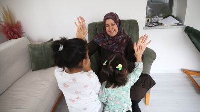 Görselin üst bölümünde, ev ortamında zeytin yeşili bir koltukta oturan başörtülü koruyucu anne ile önünde yer alan iki kız çocuğunun neşeyle el çırpma oyunu oynadığı anlar yer almaktadır. Kadın gülümseyerek çocuklara ellerini uzatırken, arkaları kameraya dönük olan çocuklar ellerini kadının elleriyle buluşturarak keyifli bir etkileşim kurmaktadır.