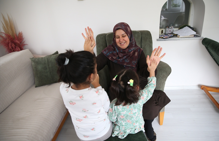Görselin üst bölümünde, ev ortamında zeytin yeşili bir koltukta oturan başörtülü koruyucu anne ile önünde yer alan iki kız çocuğunun neşeyle el çırpma oyunu oynadığı anlar yer almaktadır. Kadın gülümseyerek çocuklara ellerini uzatırken, arkaları kameraya dönük olan çocuklar ellerini kadının elleriyle buluşturarak keyifli bir etkileşim kurmaktadır.