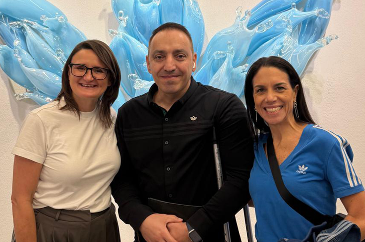 Duvar boyunca uzanan parlak mavi tonlardaki büyük adidas sanat enstalasyonunun önünde üç kişi yan yana duruyor. Solda beyaz gömlekli Michelle Robertson, ortada siyah gömleği ile Mehmet Kızıltaş, sağda mavi adidas üstüyle Ana Peniche yer alıyor. Üçlü, mağaza içindeki modern ve aydınlık atmosferde kameraya dönük şekilde poz veriyor.